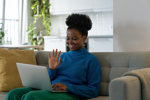 Alegre jovem afro-americana com sorriso fazendo videochamada sentada no sofá com laptop no colo