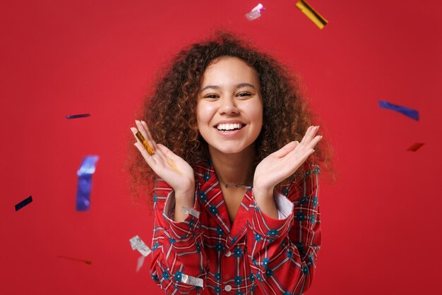 Foto alegre jovem afro-americana em homewear de pijama posando enquanto descansava em casa isolada em fundo vermelho. relaxe o conceito de estilo de vida de bom humor. mock up espaço de cópia. comemorando vomitar confete.