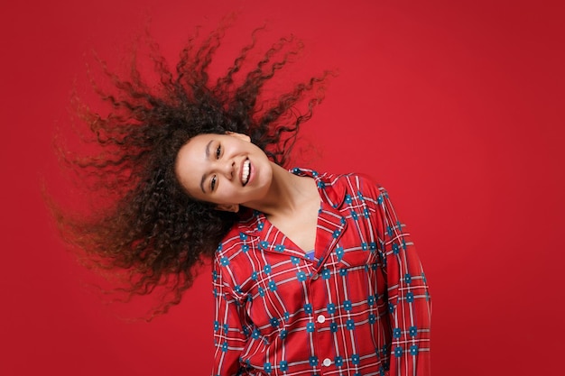 Alegre jovem afro-americana em pijama homewear posando descansando em casa isolada em fundo vermelho. Relaxe o conceito de estilo de vida de bom humor. Mock up espaço de cópia. Divertindo-se pular com o cabelo esvoaçante.