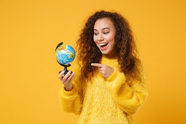 Foto alegre jovem afro-americana em suéter de pele posando isolado em fundo amarelo alaranjado, retrato de estúdio. conceito de estilo de vida de pessoas. mock up espaço de cópia. apontando o dedo indicador no globo do mundo.