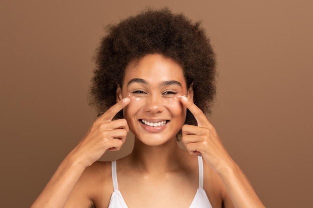 Foto alegre jovem afro-americana encaracolada com pele perfeita em top branco aplique creme no rosto sob os olhos