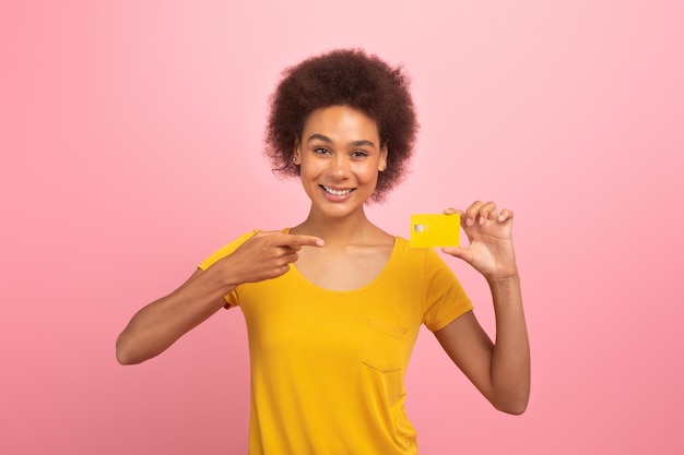 Alegre jovem afro-americana encaracolada viciada em compras em camiseta amarela aponta o dedo para o cartão de crédito