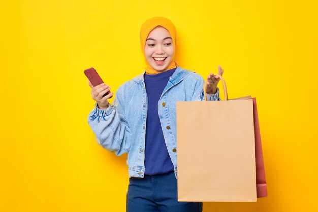 Alegre jovem asiática em jaqueta jeans segurando sacolas de compras e cartão de crédito isolado sobre fundo amarelo