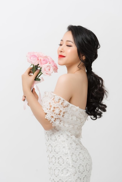 Alegre jovem asiática vestindo vestido branco e segurando buquê de flores sobre fundo branco