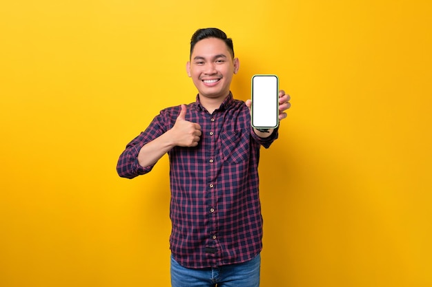 Alegre jovem asiático demonstrando smartphone com tela vazia mostrando polegar para cima gesto isolado em fundo amarelo Publicidade novo conceito de maquete de aplicativo móvel
