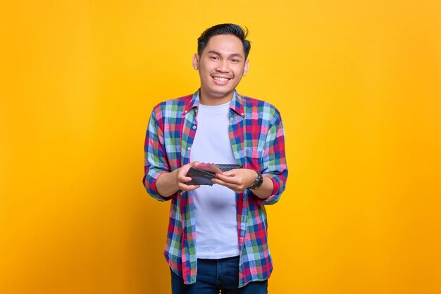 Alegre jovem asiático em camisa xadrez mostrando carteira com dinheiro isolado em fundo amarelo