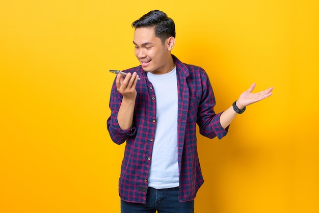 Alegre jovem asiático em camisa xadrez usando o assistente de voz no smartphone isolado em fundo amarelo