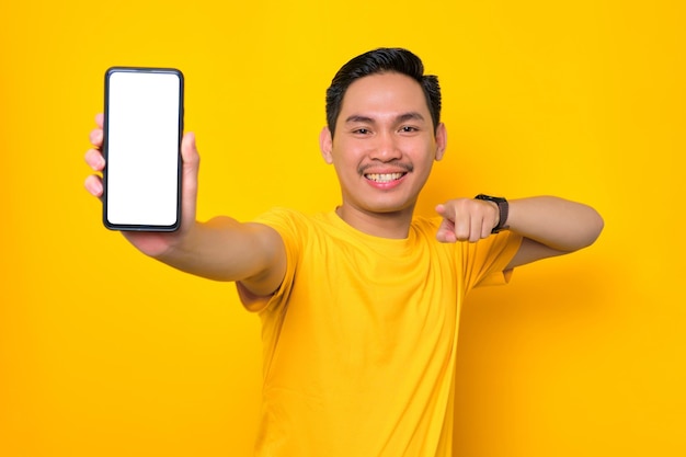 Alegre jovem asiático em camiseta casual apontando para o celular de tela em branco isolado em fundo amarelo Conceito de estilo de vida de pessoas