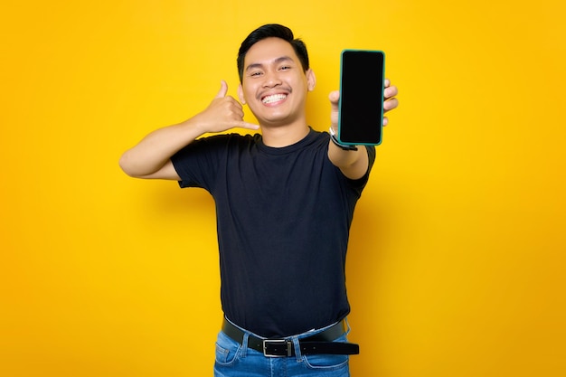 Alegre jovem asiático em camiseta casual mostrando telefone celular com tela em branco mostrando me chame gesto isolado em fundo amarelo Conceito de estilo de vida de pessoas
