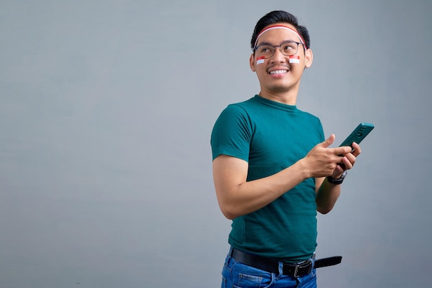 Alegre jovem asiático em camiseta casual segurando o celular e olhando de lado para o espaço de cópia isolado no conceito de celebração do dia da independência indonésia de fundo cinza