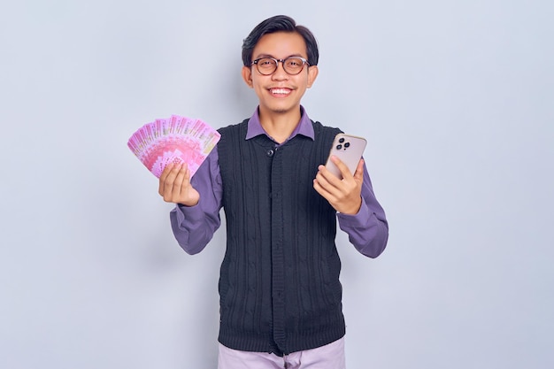 Alegre jovem asiático em roupas de camisa roxa segurando dinheiro em notas de rupia e telefone celular isolado no fundo branco Conceito de estilo de vida de pessoas