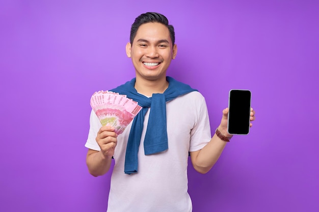 Alegre jovem asiático segurando o telefone celular com tela em branco e notas de dinheiro isoladas sobre fundo roxo Conceito de transferência de dinheiro on-line fácil