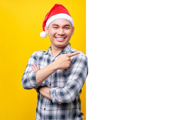 Alegre jovem asiático usando um chapéu de Natal em pé e apontando o dedo para fora do lado de uma placa de propaganda branca isolada em fundo amarelo Feliz Ano Novo 2023 conceito de feriado