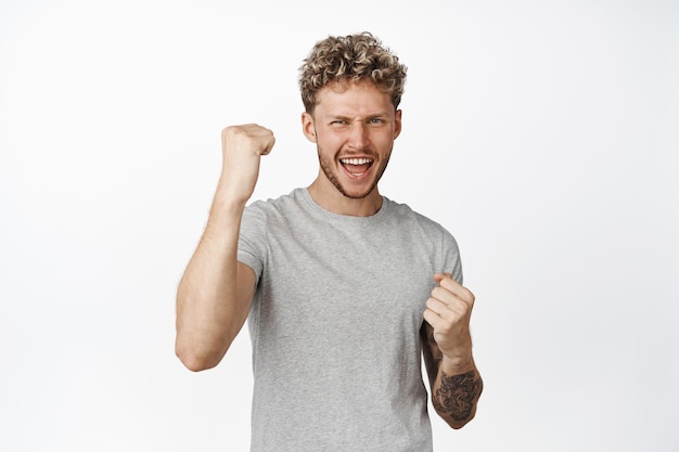 Alegre jovem bonito em camiseta cinza comemorando grito de alegria e fazendo punho bombeia objetivo alcançado e triunfando fundo branco