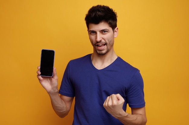 Alegre jovem bonito olhando para a câmera mostrando o celular fazendo sim gesto em fundo amarelo