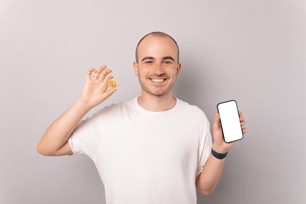 Alegre jovem careca está mostrando um bitcoin e uma tela do telefone