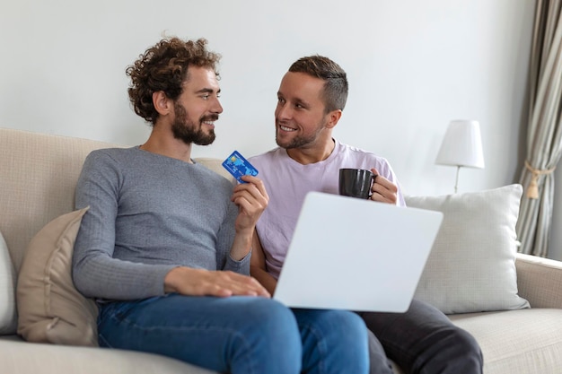 Alegre jovem casal gay sorrindo alegremente enquanto faz compras online em casa Dois jovens amantes do sexo masculino usando um cartão de crédito e um laptop para fazer uma compra online Jovem casal gay sentados juntos dentro de casa