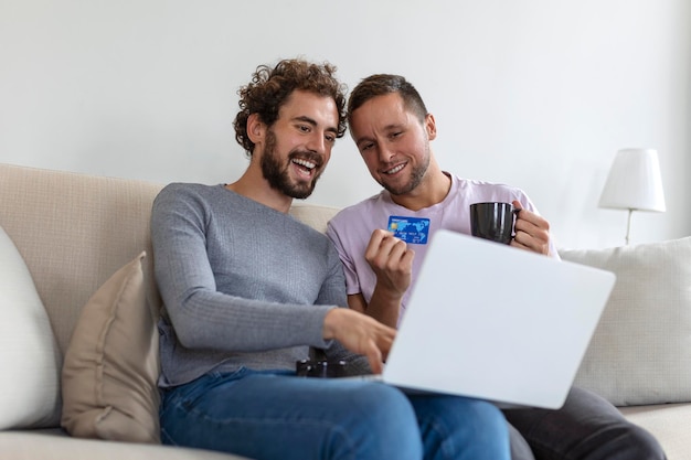Alegre jovem casal gay sorrindo alegremente enquanto faz compras online em casa Dois jovens amantes do sexo masculino usando um cartão de crédito e um laptop para fazer uma compra online Jovem casal gay sentados juntos dentro de casa