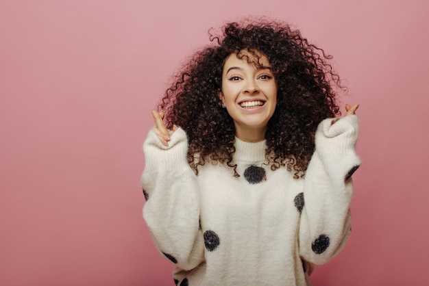 Alegre jovem caucasiana faz desejo com os dedos cruzados no fundo rosa Mulher morena de cabelos encaracolados na camisola sorri com os dentes Esperança para o conceito de boa sorte