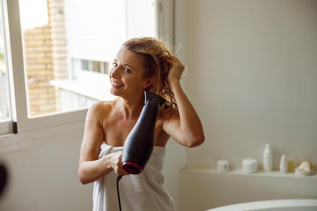 Foto alegre jovem caucasiana linda mulher secando o cabelo com secador depois de tomar banho