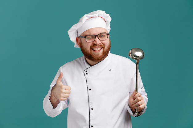 Alegre jovem chef masculino vestindo óculos uniforme e boné mostrando concha e polegar para cima olhando para câmera piscando isolada em fundo azul