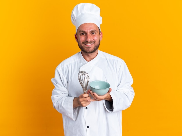 Alegre jovem cozinheiro masculino caucasiano com uniforme de chef e boné segurando a tigela e o batedor, olhando para a câmera isolada na parede laranja