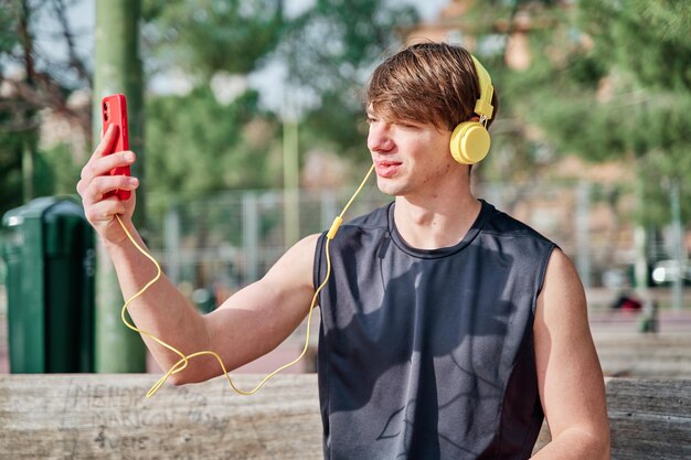 Alegre jovem desportista fazendo videochamada em seu smartphone com fone de ouvido jovem apto macho tirando uma selfie após o exercício