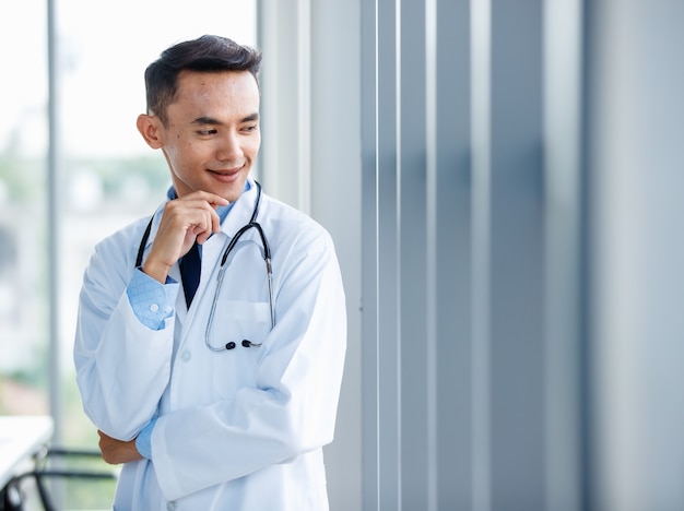Alegre jovem e bonito médico asiático masculino de uniforme e com estetoscópio na clínica e pose com gesto positivo e autoconfiante.