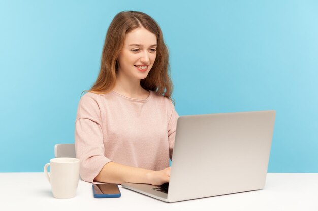 Alegre jovem empresária em roupas casuais, sentado no local de trabalho digitando no laptop e sorrindo trabalhando on-line fazendo trabalho freelance no estúdio interno do escritório em casa isolado em fundo azul