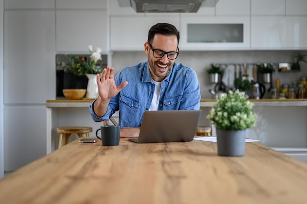 Alegre jovem empresário acenando com a mão durante uma chamada de vídeo sobre laptop na mesa no escritório em casa