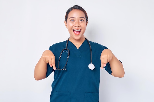 Alegre jovem enfermeira profissional asiática trabalhando vestindo um uniforme azul, apontando os dedos para baixo, mostrando um lugar livre para propaganda isolada no fundo branco Conceito de medicina de saúde