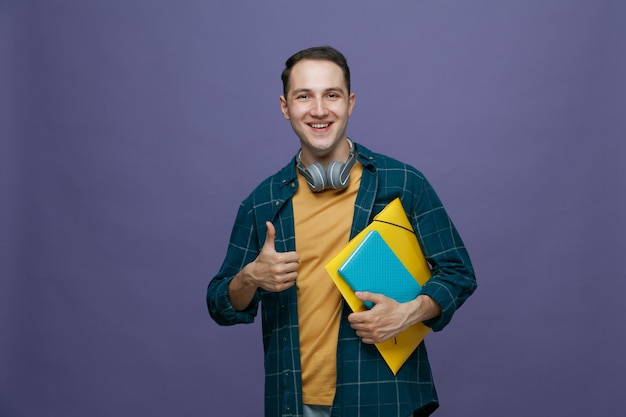Alegre jovem estudante do sexo masculino usando fones de ouvido no pescoço segurando a pasta e o caderno olhando para a câmera mostrando o polegar isolado no fundo roxo