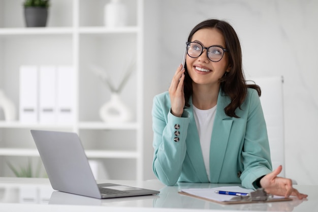 Alegre jovem europeia de óculos e terno na mesa com laptop ligando por telefone