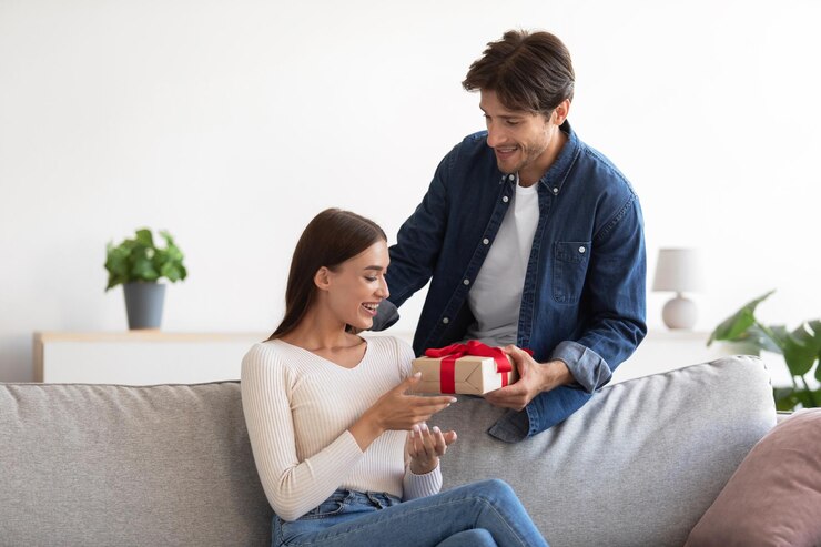 Foto alegre jovem europeu dando caixa de presente para surpreender a mulher no sofá no interior da sala de estar