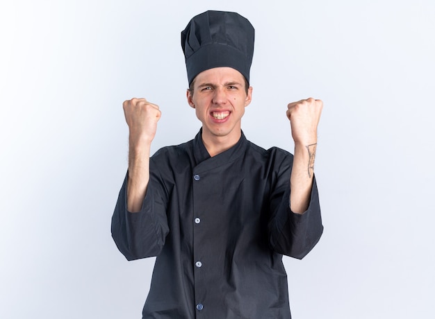 Alegre jovem loira cozinheiro masculino com uniforme de chef e boné olhando para a câmera fazendo gesto de sim isolado na parede branca