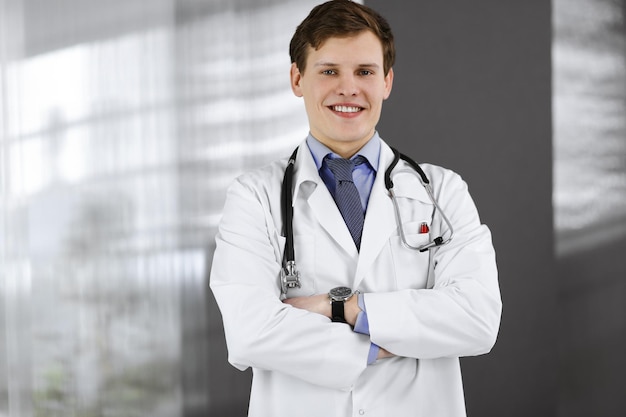 Alegre jovem médico está de pé com os braços cruzados em uma clínica. Retrato de médico profissional no trabalho. Conceito de medicina.