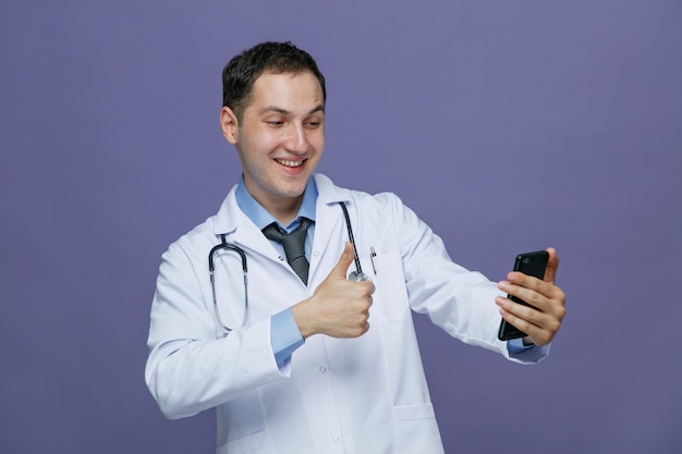 Alegre jovem médico masculino vestindo roupão médico e estetoscópio em volta do pescoço esticando o celular mostrando o polegar para cima falando via videochamada isolada no fundo roxo