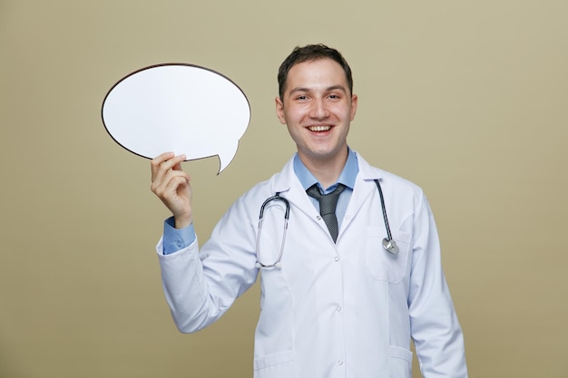 Alegre jovem médico masculino vestindo roupão médico e estetoscópio no pescoço, olhando para a câmera mostrando a bolha de bate-papo isolada no fundo verde-oliva