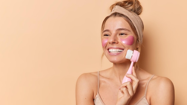 Alegre jovem modelo feminino europeu usa sorrisos de massageador de rosto aplica manchas de hidrogel rosa sob os olhos desvia o olhar usa faixa de cabeça fica ombros nus isolados sobre fundo marrom