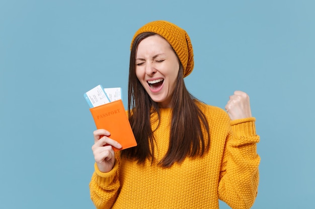 Alegre jovem morena de chapéu de suéter posando isolado no retrato de estúdio de fundo azul. Conceito de estilo de vida de pessoas. Mock up espaço de cópia. Segure os bilhetes de embarque do passaporte fazendo o gesto do vencedor.