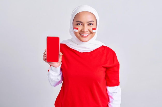 Alegre jovem muçulmana asiática em camiseta branca vermelha mostrando a tela em branco do celular recomendando App isolado no fundo branco comemorando o dia da independência indonésia em 17 de agosto