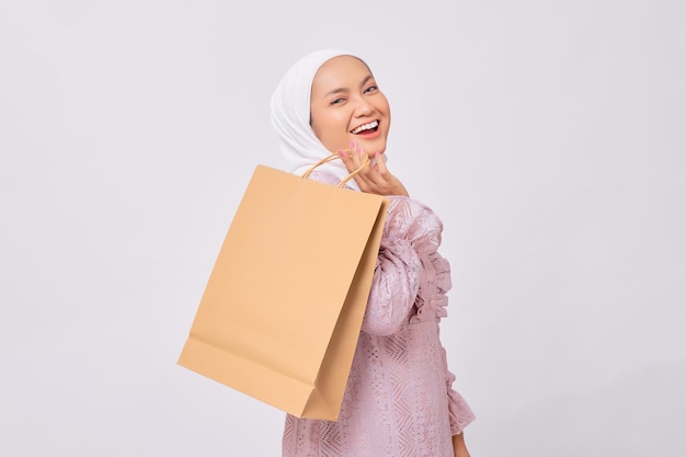 Alegre jovem muçulmana asiática usando hijab e vestido roxo segurando sacola de compras isolada no fundo branco do estúdio