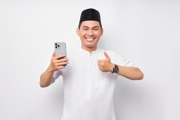 Alegre jovem muçulmano asiático segurando smartphone e mostrando o polegar para cima gesto isolado no fundo branco Celebração do conceito de estilo de vida do Islã religioso pessoas Ramadã e Ied Mubarak