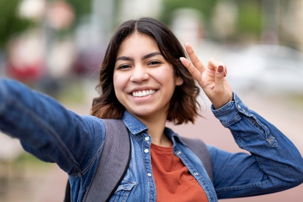 Alegre jovem mulher árabe tirando selfie ao ar livre mostrando gesto de paz para a câmera