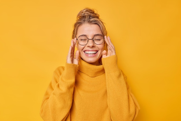 Alegre jovem mulher caucasiana mantém os olhos fechados sorrisos dentuço usa jumper casual mantém as mãos na borda dos óculos tem bom humor isolado sobre fundo amarelo vívido. Conceito de emoções felizes