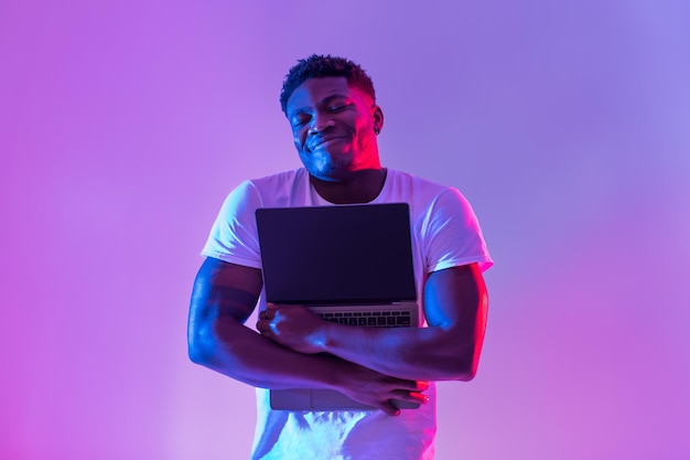 Foto alegre jovem negro abraçando laptop com tela vazia em maquete de luz neon para site