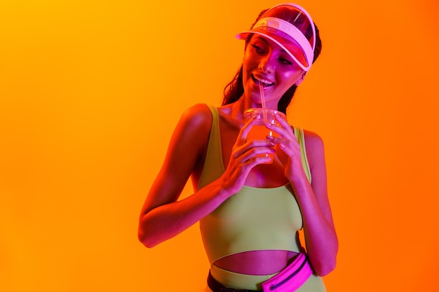 alegre jovem otimista elegante em roupas de estilo de vida do esporte e tampa transparente, bebendo um coquetel de suco isolado sobre uma parede laranja com luzes de néon.