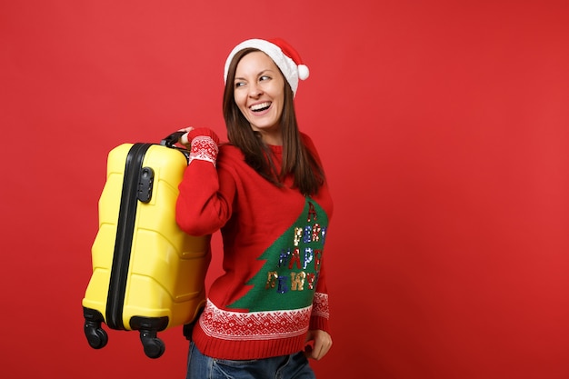 Alegre jovem santa na camisola de malha, chapéu de Natal, olhando para trás, segurando a mala amarela isolada sobre fundo vermelho. Conceito de festa de feriado de celebração de ano novo feliz 2019. Simule o espaço da cópia.