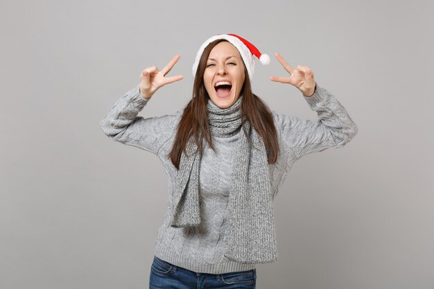 Alegre jovem santa no chapéu de Natal do cachecol de camisola cinza mostrando sinal de vitória isolado no fundo da parede cinza no estúdio. Conceito de festa de feriado de celebração de ano novo feliz 2019. Simule o espaço da cópia.