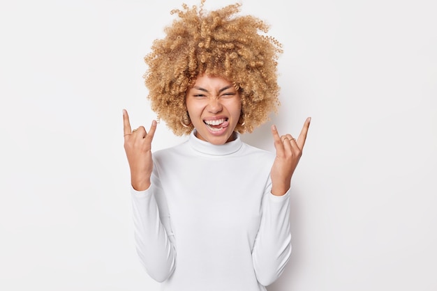 Alegre jovem sortuda com cabelo encaracolado mostra gesto de rock n roll assiste a um show de festa incrível faz sinal de heavy metal sobressai a língua usa gola alta casual isolada sobre fundo branco